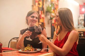 Two young women smiling and clinking their wine glasses together at a restaurant.