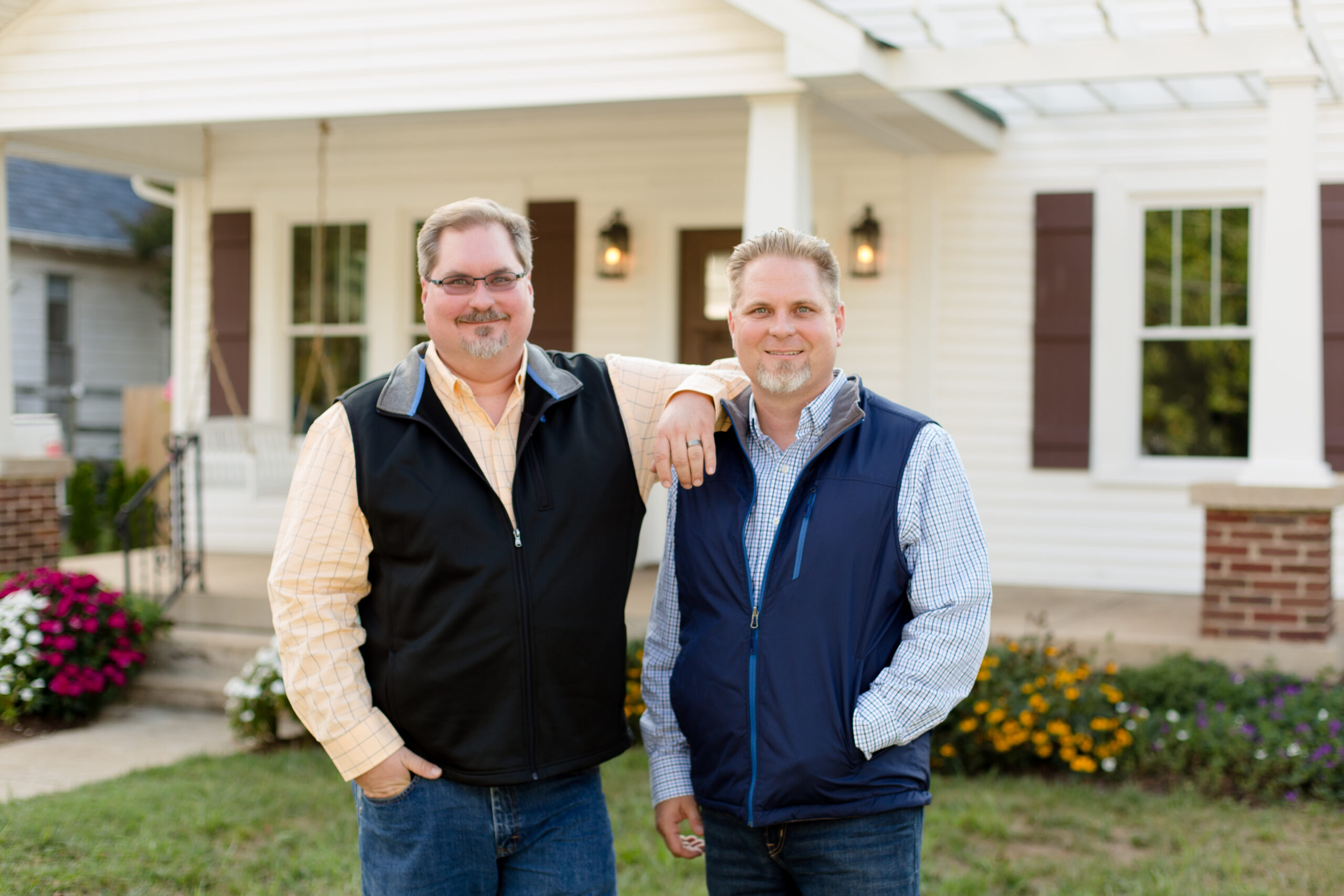 Two men stand with arms around each other in a front yard of a beige house with white trim and flowers nearby, smiling at the camera.